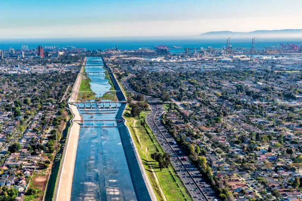 The levees and flood control channel making up the Los Angeles River shot facing westerly with the skyline of Long Beach and the Port of Los Angeles in the distance from over the cities of Long Beach and Carson, California.