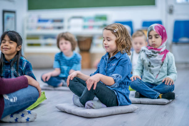 School Aged Children Meditating in Class A diverse group of young school children are indoors, sitting cross legged on their classroom floor. They are each sitting on their pillows with their hands on their knees and eyes closed as they focus on meditating. mindfulness kids stock pictures, royalty-free photos & images