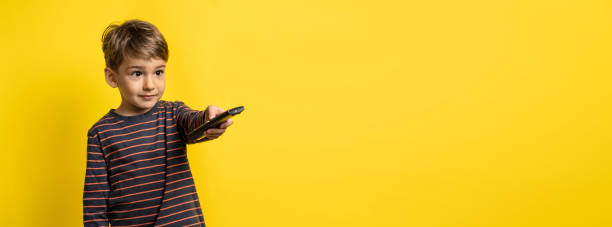 SMall caucasian boy holding tv remote controller while standing in front of yellow background changing channel with copy space