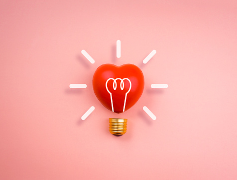 Light bulb icon on red heart ball on pink pastel background, minimal style. Love, care, sharing, giving, wellbeing, inspiration, and idea concepts.