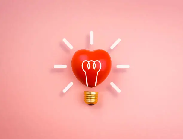 Photo of Light bulb icon on red heart ball on pink pastel background, minimal style. Love, care, sharing, giving, wellbeing, inspiration, and idea concepts.