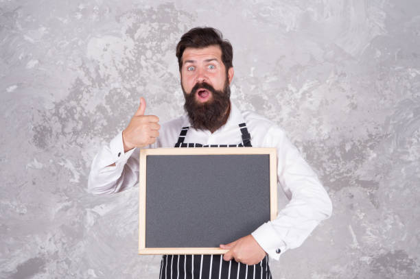 istock culinary school classes. successful cook career. best cuisine here. welcome on board. brutal man hipster hold blackboard, copy space. nicest price in town. surprised bearded chef show thumb up 1359953352