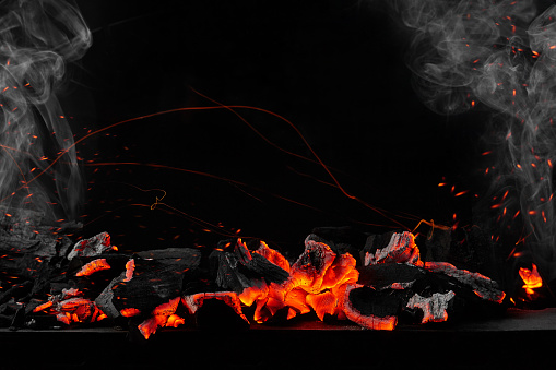 Red hot burning coals on metal barbecue tray against black background with soaring bright sparks and light white smoke. Horizontal image