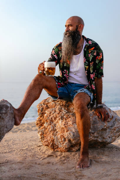 careca cabeça homem barbudo vestindo com camisa havaiana relaxado enquanto sentado em uma grande pedra de uma praia e bebendo uma cerveja. - beer glass mustache beer color image - fotografias e filmes do acervo