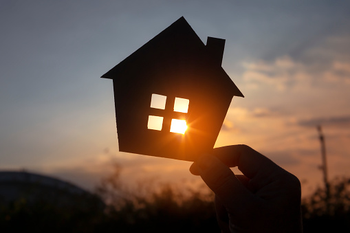 Hands holding paper house on sunset sky, family home, homeless housing, and home protection insurance concept, international day of families, new life, foster home care, and real estate.