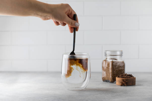 Woman stirs instant coffee in glass mug with boiled water on grey stone table Woman stirs instant granulated coffee in glass mug with hot boiled water by spoon on grey stone table in light kitchen closeup glass cup of boiling water stock pictures, royalty-free photos & images
