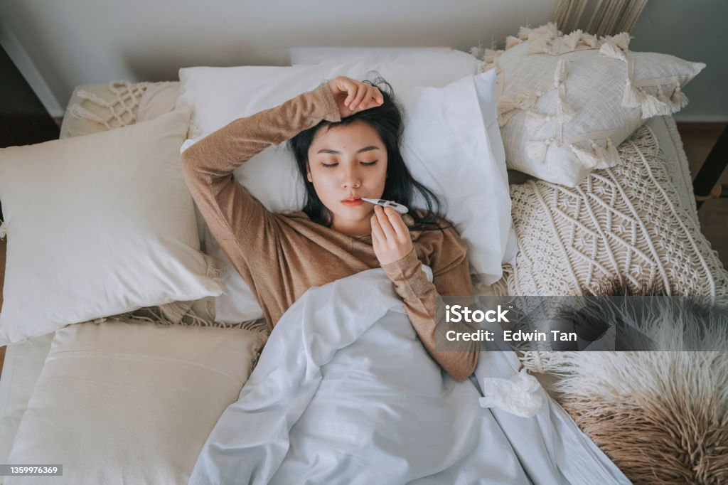directly above Asian Chinese Woman lying on bed covered with blanket having  cold and flu measured her body temperature with digital thermometer Illness Stock Photo