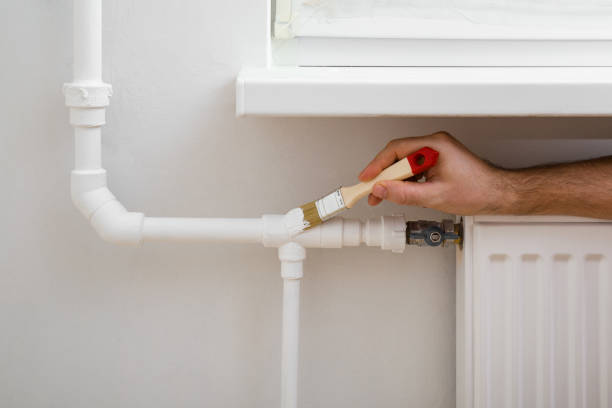 Young adult man hands using brush and painting heating pipe white color. Closeup. Repair work of home. stock photo