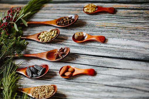 Close up of typical tasty Christmas and winter spices in nine dark wooden spoons on rustic wooden grunge background. The spices are and tonka bean, coriander, anise star, cardamom, almond, clove, walnut, hazelnut, nutmeg, cinnamon and with christmas decoration on fir branch. Copy space. Selective focus and color editing with added grain. Part of a series.