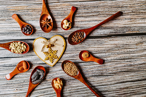 Beautiful flat lay of delicious Christmas spices in nine dark wooden spoons arranged to a poinsettia with a heart on rustic wooden grunge background with some copy space. The spices are Tonka bean, juniper berry, coriander, anise star, cardamom, almond, clove, walnut, hazelnut, nutmeg and cinnamon. Copy space. Color editing with added grain. Part of a series.