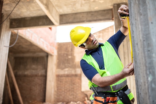 Male worker measuring a wall with measure tape at construction site. Man wearing yellow vest and hardhat working at building site.