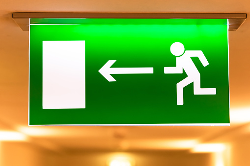 A bright green emergency evacuation sign with a directional arrow and a running figure hanging from the ceiling in the hallway. Selective focus. Close-up