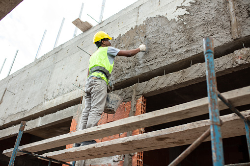Low angle view of a mason worker plastering exterior building wall on construction site. Male worker standing on scaffolding putting on cement plaster on wall.