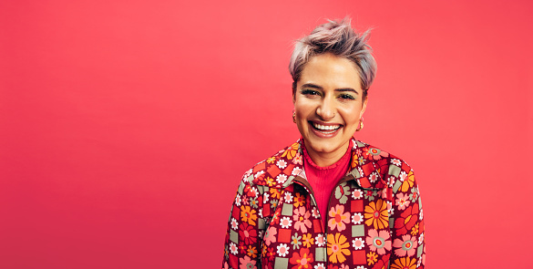 Hipster woman looking at the camera with a happy smile on her face. Cheerful young woman standing alone against a red background. Fashionable young woman with dyed hair feeling vibrant.