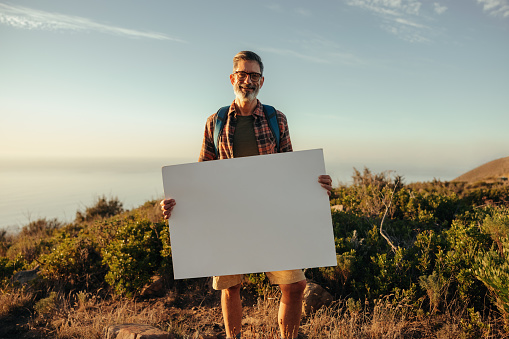 Hiker holding a blank placard on a hilltop. Happy mature backpacker displaying a banner while standing on top of a hill at sunset. Adventurous male activist smiling at the camera.