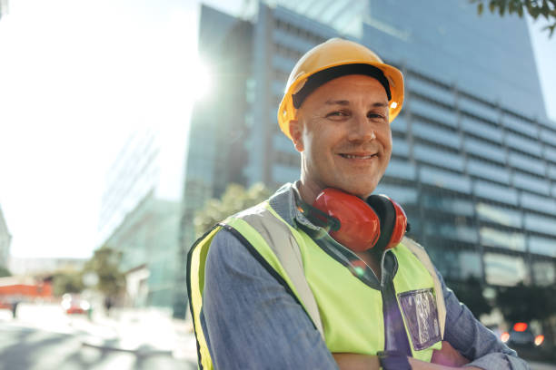 Happy construction worker smiling at the camera in the city Happy construction worker smiling at the camera while standing with his arms crossed in the city. Mid-adult blue collar worker standing in front of high rise buildings in his workwear. blue collar worker stock pictures, royalty-free photos & images