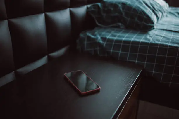 Comfortable bed and nightstand with black smartphone in modern hotel room interior