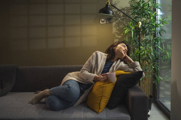 istock Young woman sitting on sofa and drinking tea while looking through window 1360007640