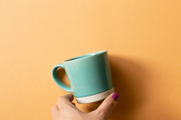 istock Hand holding mint green mug cup on orange background. top view, copy space 1360014122