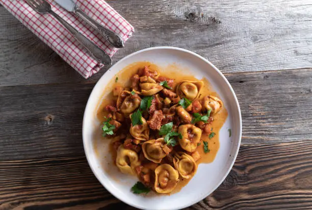 Homemade rustic pasta dish with fresh tortellini with pancetta, tomatoes and vegetable sauce. Served isolated on wooden table from above.