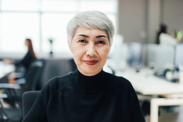 Portrait of Asian senior female manager at desk in the office looking at camera. POV of meeting at video conference. Portrait of Asian senior female manager at desk in the office looking at camera. POV of meeting at video conference. headshot stock pictures, royalty-free photos & images