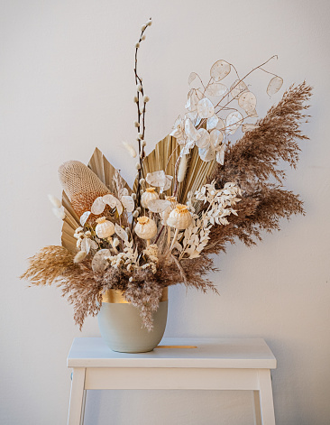 Beautiful flower arrangement with dry plants - lunaria branches, poppy, pampas and others in a handmade pot standing on a wooden table over a white wall.