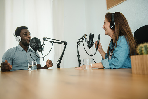 Cheerful diverse coworkers in headphones sitting at table and speaking in mics while recording podcast together in studio