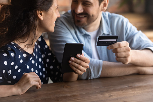 Cropped happy young family couple holding telephone and credit bank card, enjoying shopping in internet store, paying for goods, purchasing services online, booking hotel or tickets, transfer money.