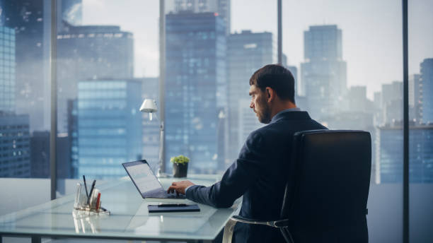 istock Relaxed Businessman in a Suit Sitting at a Desk in Modern Office, Using Laptop Computer, Next to Window with Big City with Skyscrapers View. Successful Finance Manager Planning Work Projects. 1360028388