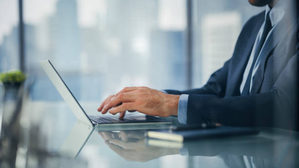 istock Close Up on Businessman Sitting at a Desk in Modern Office, Using Laptop Computer, Next to Window with Big City with Skyscrapers View. Successful Finance Manager Planning Work Projects. 1360028402