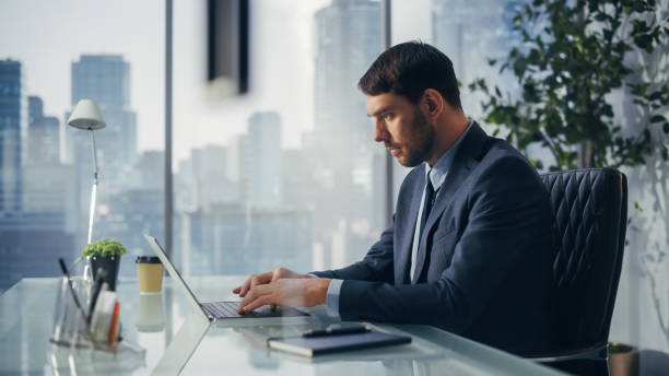 istock Confident Businessman in a Suit Sitting at a Desk in Modern Office, Using Laptop Computer, Next to Window with Big City with Skyscrapers View. Successful Finance Manager Planning Work Projects. 1360028405