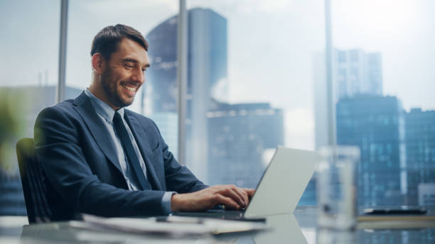 istock Smiling Businessman in a Suit Sitting at a Desk in Modern Office, Using Laptop Computer, Next to Window with Big City with Skyscrapers View. Successful Finance Manager Planning Work Projects. 1360028492