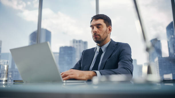 istock Businessman in a Suit Sitting at a Desk in Modern Office, Using Laptop Computer, Next to Window with Big City with Skyscrapers View. Successful Finance Manager Resting with Closed Eyes. 1360028541