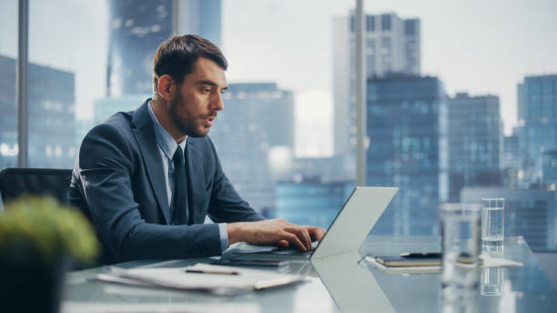istock Happy Businessman in a Suit Sitting at a Desk in Modern Office, Using Laptop Computer, Next to Window with Big City with Skyscrapers View. Successful Finance Manager Planning Work Projects. 1360028581