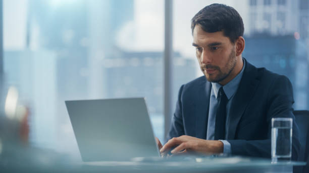 istock Confident Businessman in a Suit Sitting at a Desk in Modern Office, Using Laptop Computer, Next to Window with Big City with Skyscrapers View. Successful Finance Manager Planning Work Projects. 1360028623