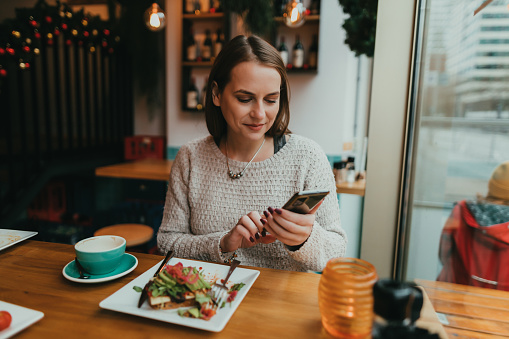 Young woman sending text messages on a smartphone and eating avocado toast for breakfast in a restaurant