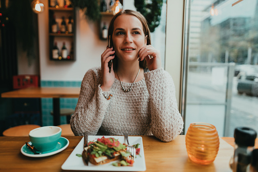 Young woman sitting in a restaurant and talking on a smartphone, eating avocado toast for breakfast
