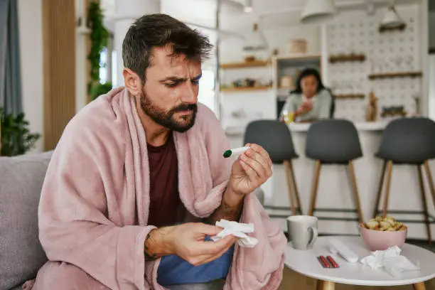 Bearded sick man with flue sitting on sofa at home and measuring body temperature.