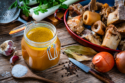 High angle view of healthy homemade bone broth in an open airtight glass container shot on rustic wooden table. Ingredients for cooking bone broth are all around the container. High resolution 42Mp studio digital capture taken with Sony A7rII and Sony FE 90mm f2.8 macro G OSS lens