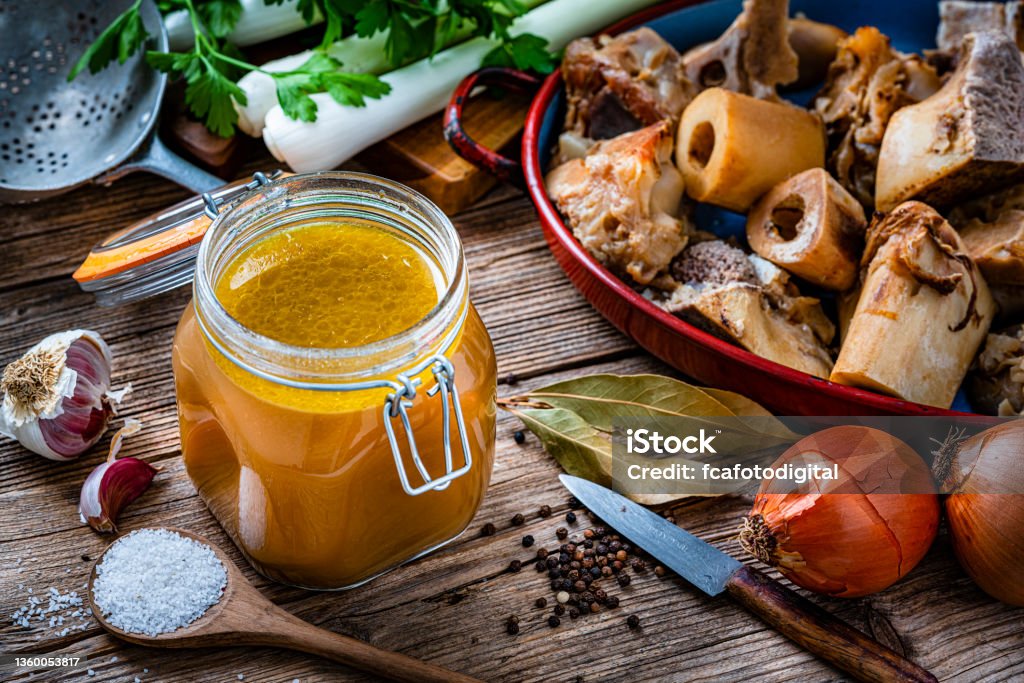 Bone broth and ingredients on rustic table High angle view of healthy homemade bone broth in an open airtight glass container shot on rustic wooden table. Ingredients for cooking bone broth are all around the container. High resolution 42Mp studio digital capture taken with Sony A7rII and Sony FE 90mm f2.8 macro G OSS lens Bone Broth Stock Photo