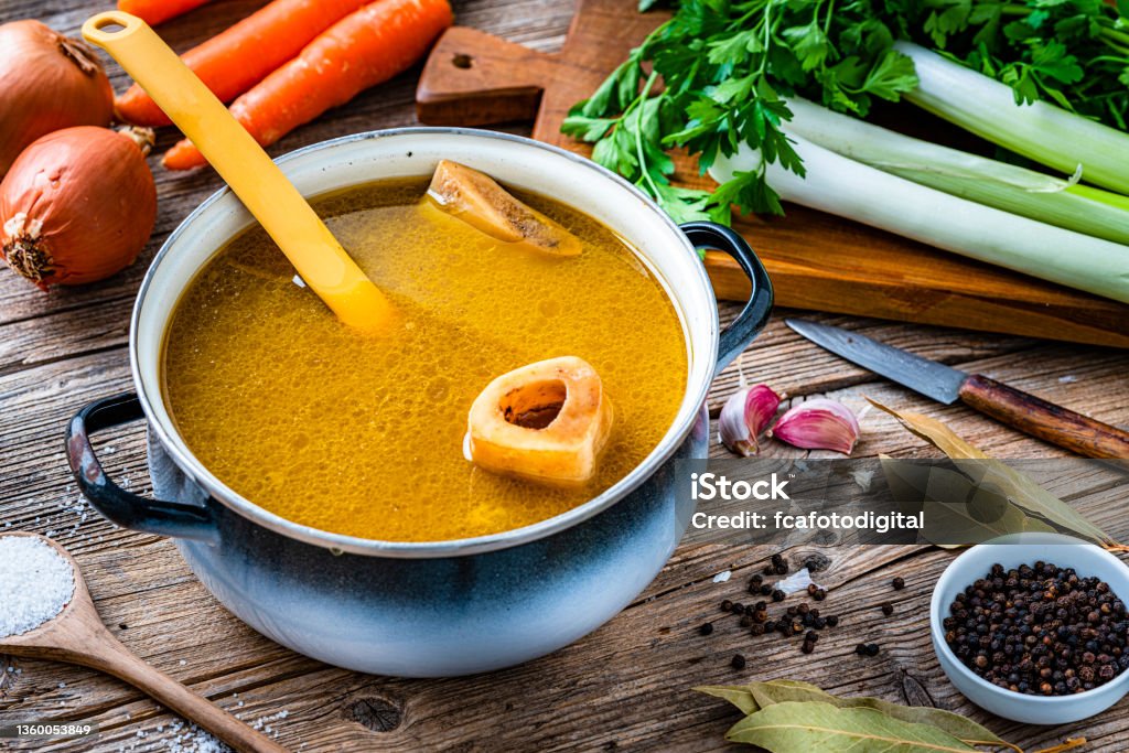 Bone broth in a cooking pan High angle view of a cooking pan filled with homemade bone broth shot on rustic wooden table. Ingredients for cooking bone broth are all around the pan. High resolution 42Mp studio digital capture taken with Sony A7rII and Sony FE 90mm f2.8 macro G OSS lens Bone Broth Stock Photo