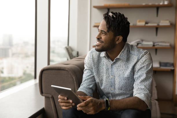 istock Smiling African guy holding digital tablet looking out window 1360060891