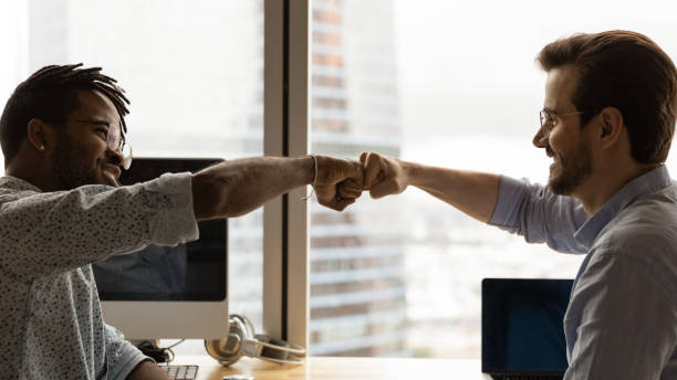 Diverse male colleagues giving fist bump at workplace Diverse male colleagues giving fist bump at workplace. African guy greeting Caucasian mate, symbol of business partnership, racial equality, friendship at work. Respect, support, collaboration concept male friendship stock pictures, royalty-free photos & images