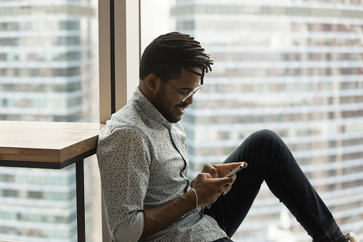 Handsome hipster African guy in glasses sit in modern apartment, take break in office space with smartphone, urban skyscrapers city view through window. Lifestyle, modern tech usage, business concept