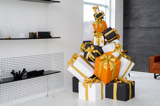 Christmas present gift box stacked on floor in living room white interior. Holiday sale concept. Many gift box black, white, orange color with gold ribbon