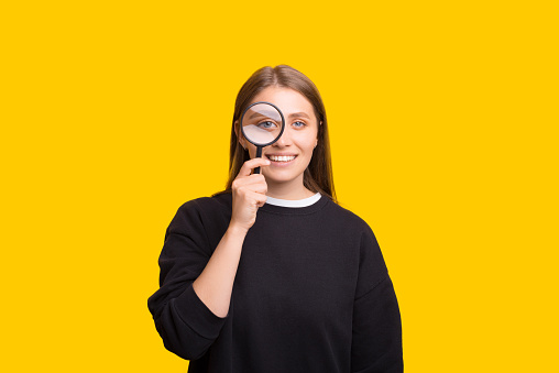 Portrait of cheerful pretty woman looking at camera through magnifying glass, isolated over yellow background