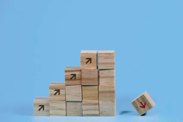 Photo of Wooden cubes build as stair steps with arrows pointing upwards, but the last cube is falling down, concept for the risk of failure due to overly rapid business growth, blue background with copy space