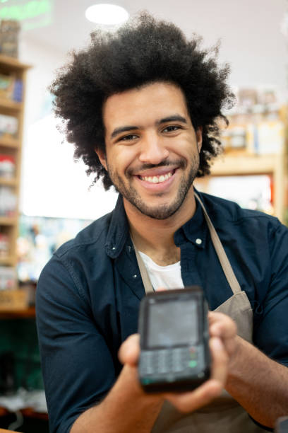 istock Cheerful black man working at a supermarket holding a credit card reader while facing camera smiling very happy 1360082596