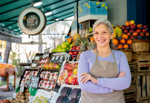 Portrait of Latin American senior business owner of a fruit and vegetable stand wearing an apron facing camera smiling with arms crossed - Small food business concepts