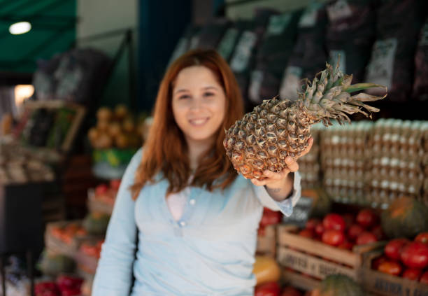Latin American woman at a fruit and vegetable stall holding a pineapple while facing camera smiling - Focus on foreground